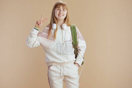 Photo for Portrait of smiling trendy young woman in beige tracksuit with backpack and headphones isolated on beige showing victory gesture. - Royalty Free Image