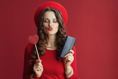 Téléchargez les photos : Joyeux Valentin. moderne 40 ans femme en robe rouge et béret isolé sur fond rouge avec smartphone et bonbons en forme de coeur sur bâton envoi d'un baiser. - en image libre de droit
