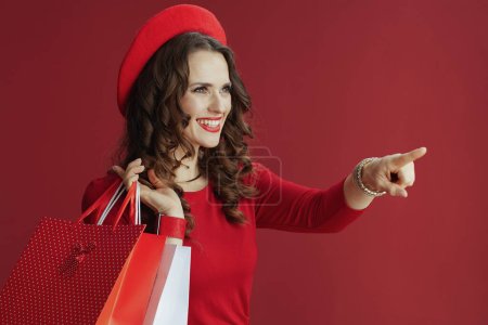 Photo for Happy Valentine. happy stylish middle aged woman in red dress and beret pointing at something isolated on red background with shopping bags. - Royalty Free Image