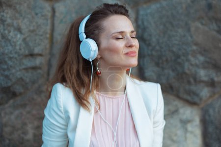 Photo for Elegant middle aged woman in pink dress and white jacket in the city with headphones against stone wall. - Royalty Free Image