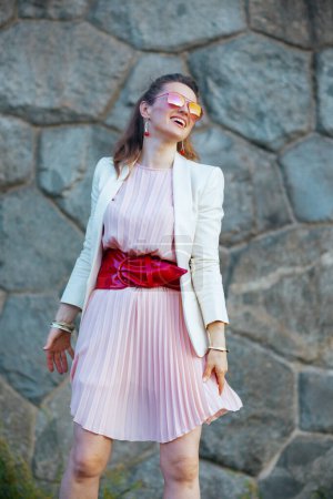 Téléchargez les photos : Heureuse femme d'âge moyen élégant en robe rose et veste blanche dans la ville avec des lunettes de soleil contre le mur de pierre. - en image libre de droit