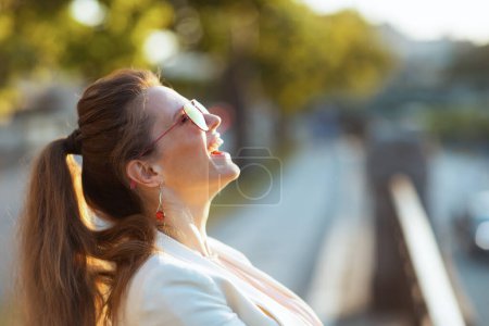 Foto de Mujer elegante sonriente en vestido rosa y chaqueta blanca en la ciudad con gafas de sol. - Imagen libre de derechos