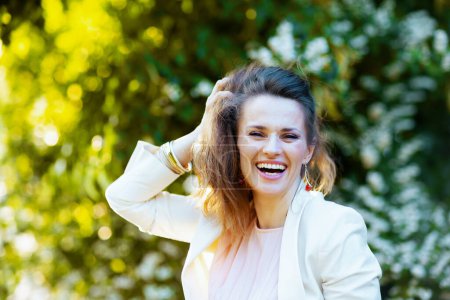 Téléchargez les photos : Portrait de heureuse femme d'âge moyen élégant en robe rose et veste blanche dans la ville contre la verdure. - en image libre de droit
