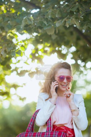 Téléchargez les photos : Heureuse jeune femme en robe rose et veste blanche dans la ville avec sac rouge et lunettes de soleil à l'aide d'un smartphone. - en image libre de droit