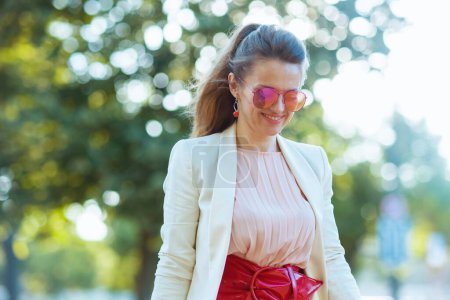 Téléchargez les photos : Heureuse femme d'âge moyen moderne en robe rose et veste blanche dans la ville avec des lunettes de soleil marche. - en image libre de droit