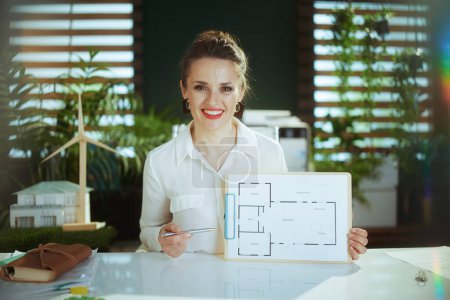 Photo for Sustainable real estate business. smiling young female realtor in modern green office in white blouse with clipboard and building plan. - Royalty Free Image
