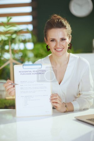 Foto de Negocio inmobiliario sostenible. sonriente mujer elegante agente de bienes raíces en moderna oficina verde en blusa blanca con portapapeles y documento. - Imagen libre de derechos