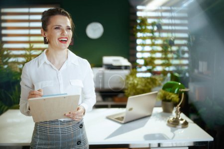 Foto de Negocio inmobiliario sostenible. mujer de negocios de mediana edad sonriente en moderna oficina verde en blusa blanca con portapapeles. - Imagen libre de derechos