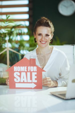 Photo for Time to move on. smiling young woman realtor in modern green office in white blouse with home for sale sign. - Royalty Free Image