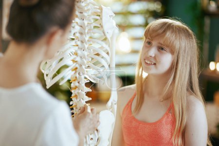 Photo for Healthcare time. smiling female massage therapist in massage cabinet with teenage client and skeleton. - Royalty Free Image