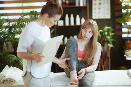Photo for Healthcare time. concerned massage therapist woman in massage cabinet with clipboard and teenage client listening to a clients complaint of leg pain. - Royalty Free Image