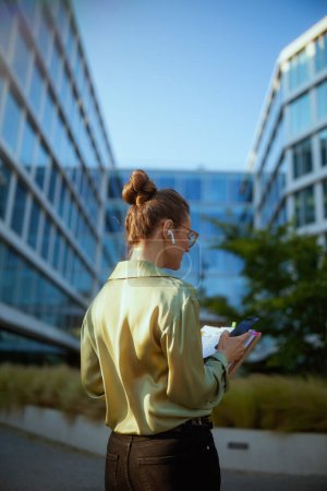 Téléchargez les photos : Vu de derrière employée près de l'immeuble de bureaux en chemisier vert avec des documents à l'aide d'un smartphone. - en image libre de droit