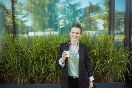 Photo for Smiling modern female employee near business center in black jacket with cup of coffee. - Royalty Free Image