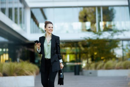 Photo for Happy modern 40 years old woman worker near business center in black jacket with briefcase using smartphone and walking. - Royalty Free Image