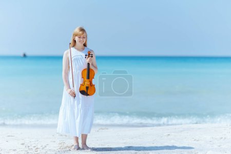Téléchargez les photos : Portrait pleine longueur d'heureuse fille moderne en robe blanche sur la côte de l'océan avec violon. - en image libre de droit