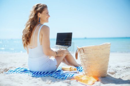 Téléchargez les photos : Heureuse femme élégante sur la plage avec sac de paille, écran blanc pour ordinateur portable et serviette rayée. - en image libre de droit
