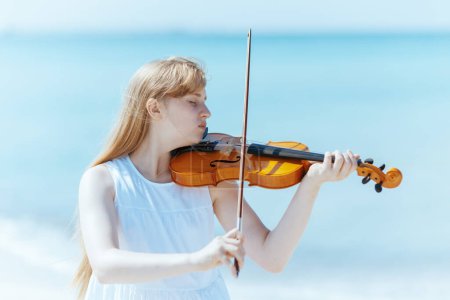Photo for Modern teenager girl in white dress with violin enjoying playing on the seacoast. - Royalty Free Image
