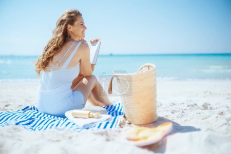 Téléchargez les photos : Femme élégante souriante sur la plage avec sac de paille, livre et serviette rayée. - en image libre de droit