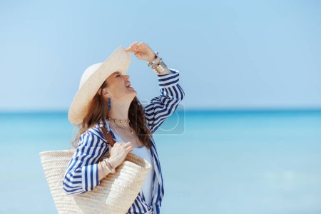 Foto de Sonriente elegante mujer de 40 años en la orilla del mar con bolsa de paja y sombrero de paja que se divierten. - Imagen libre de derechos