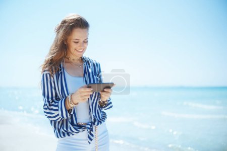 Foto de Mujer de mediana edad elegante feliz en la orilla del mar con teléfono inteligente. - Imagen libre de derechos