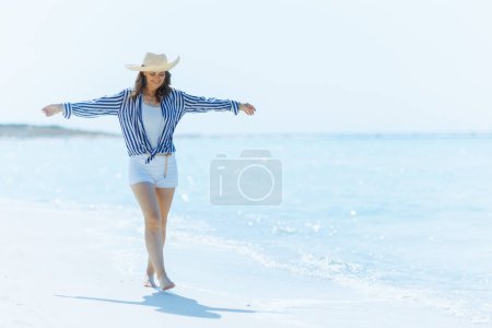 Photo for Full length portrait of smiling modern 40 years old woman on the beach with straw hat walking. - Royalty Free Image