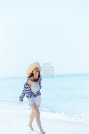 Photo for Full length portrait of happy elegant 40 years old woman on the ocean coast with straw hat walking. - Royalty Free Image