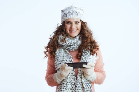 Foto de Hola invierno. sonriente mujer moderna de 40 años en suéter, mitones, sombrero y bufanda con copo de nieve utilizando aplicación de teléfono inteligente aislado en blanco. - Imagen libre de derechos