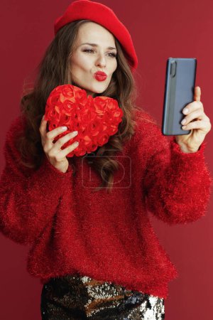 Téléchargez les photos : Joyeux Valentin. élégant 40 ans femme en pull rouge et béret avec coeur rouge et smartphone ayant webcam réunion. - en image libre de droit