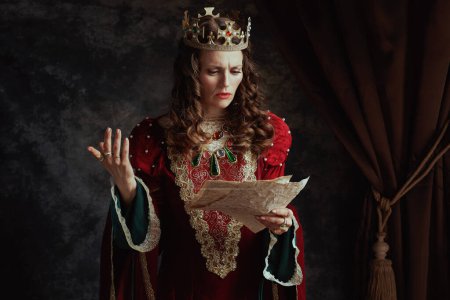 Téléchargez les photos : Malheureuse reine médiévale en robe rouge avec parchemin et couronne sur fond gris foncé. - en image libre de droit