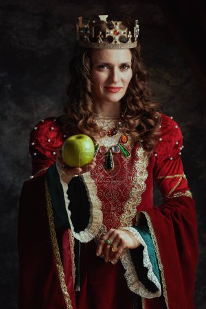 Téléchargez les photos : Reine médiévale souriante en robe rouge avec pomme verte et couronne sur fond gris foncé. - en image libre de droit