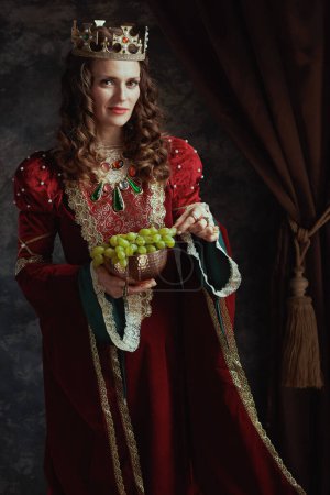 Téléchargez les photos : Reine médiévale en robe rouge avec assiette de raisins et couronne sur fond gris foncé. - en image libre de droit