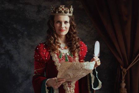 Foto de Feliz reina medieval en vestido rojo con pergamino y corona sobre fondo gris oscuro. - Imagen libre de derechos