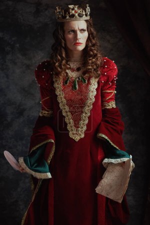 Photo for Furious medieval queen in red dress with parchment and crown on dark gray background. - Royalty Free Image