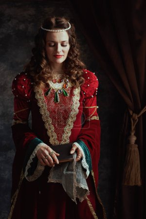 Photo for Medieval queen in red dress with book and handkerchief on dark gray background. - Royalty Free Image