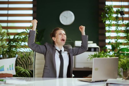 Téléchargez les photos : Un nouveau boulot. souriant moderne employée dans un bureau vert moderne en costume d'affaires gris avec ordinateur portable jetant des documents. - en image libre de droit