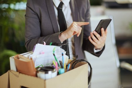 New job. Closeup on modern 40 years old woman worker in modern green office in grey business suit with personal belongings in cardboard box and smartphone.