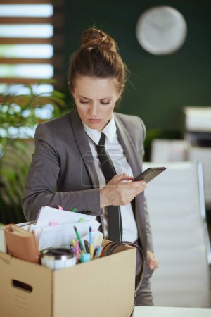 Téléchargez les photos : Un nouveau boulot. concerné femme d'âge moyen moderne employée dans un bureau vert moderne en costume d'affaires gris avec des effets personnels dans une boîte en carton à l'aide d'un smartphone. - en image libre de droit