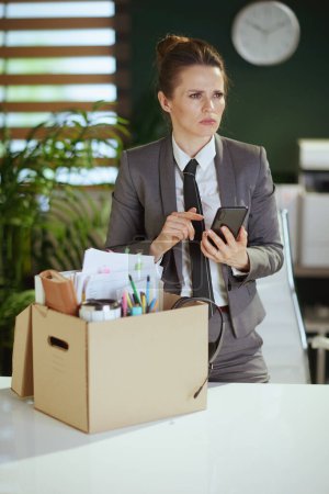 Photo for New job. pensive modern 40 years old woman worker in modern green office in grey business suit with personal belongings in cardboard box talking on a smartphone. - Royalty Free Image