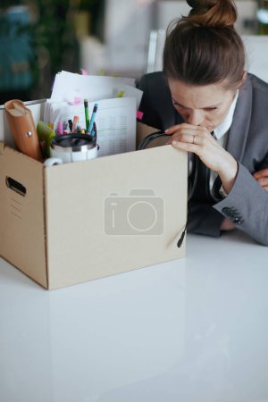 Foto de Un nuevo trabajo. infeliz mujer de mediana edad moderna trabajadora en moderna oficina verde en traje de negocios gris con pertenencias personales en caja de cartón. - Imagen libre de derechos