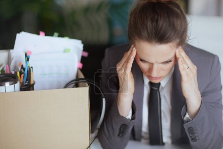 Photo for New job. pensive modern middle aged woman worker in modern green office in grey business suit with personal belongings in cardboard box. - Royalty Free Image