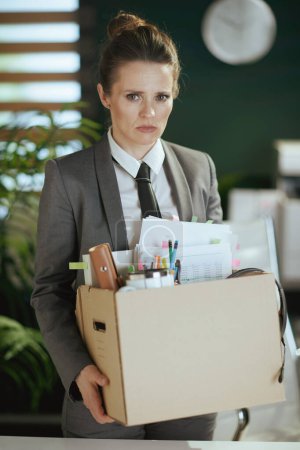 Photo for New job. sad modern middle aged woman worker in modern green office in grey business suit with personal belongings in cardboard box. - Royalty Free Image