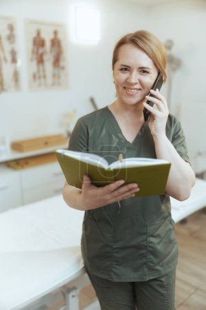 Photo for Healthcare time. happy female medical massage therapist in massage cabinet with notebook talking on a smartphone. - Royalty Free Image