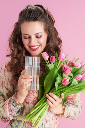 Téléchargez les photos : Jeune femme souriante aux longs cheveux bruns ondulés avec bouquet de tulipes et verre d'eau isolé sur rose. - en image libre de droit