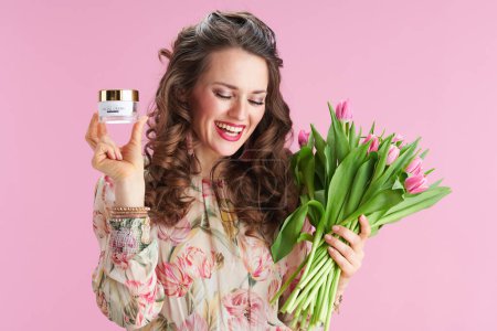 Téléchargez les photos : Heureuse femme à la mode de 40 ans avec de longs cheveux bruns ondulés avec bouquet de tulipes et crème faciale isolée sur fond rose. - en image libre de droit