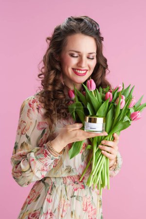 Téléchargez les photos : Jeune femme souriante en robe florale avec bouquet de tulipes et crème faciale isolée sur rose. - en image libre de droit