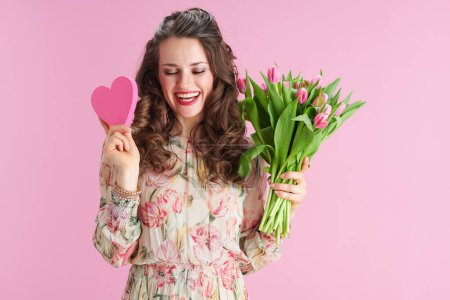 Téléchargez les photos : Heureuse femme tendance en robe florale avec bouquet de tulipes et coeur rose isolé sur rose. - en image libre de droit