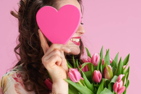 Foto de Sonriente mujer elegante con el pelo largo y ondulado morena con tulipanes ramo y corazón rosa aislado sobre fondo rosa. - Imagen libre de derechos