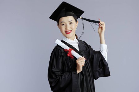 Foto de Sonriendo moderno graduado estudiante asiático mujer con diploma lanzando borlas contra gris fondo. - Imagen libre de derechos
