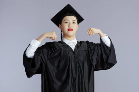 Photo for Proud modern graduate student asian woman in graduation gown with cap showing biceps against grey background. - Royalty Free Image