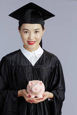 Photo for Happy young female asian graduate student with piggy bank isolated on grey background. - Royalty Free Image